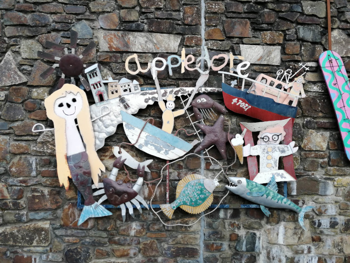Whimsical, handcrafted artwork affixed to a stone wall. The piece is a collage of repurposed metal, featuring a mermaid, boats, fish, a crab, and other sea-related imagery, along with buildings and the word Appledore prominently displayed. The style is naive and childlike, with bright colors and simple forms. The overall effect is playful and charming, suggesting a coastal or seaside location.