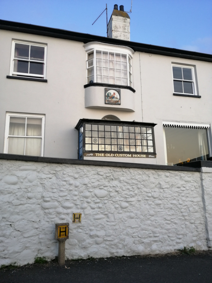 Two-story building, identified as The Old Custom House, with a whitewashed stone wall in front. The building features a bay window with a decorative tile depicting a bird, likely a rooster or similar fowl. There's also a smaller, black-framed window box below the bay window. A hydrant marker is visible on the wall. The overall style suggests an older building, possibly historic.