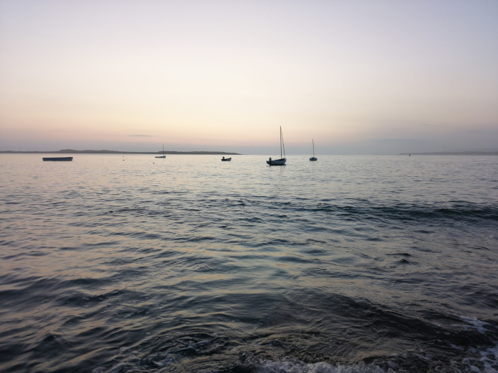 Calm sea at sunset or sunrise. A few small boats, including sailboats, are visible on the horizon. The sky is a pale, soft colour, and the overall mood is serene and peaceful.