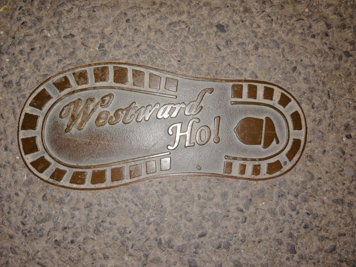 Bronze-coloured metal inlay set into a paved surface. The inlay is shaped like a footprint and contains the words Westward Ho! in a stylised script font, along with a small acorn image. The design suggests a playful reference to westward expansion or travel.