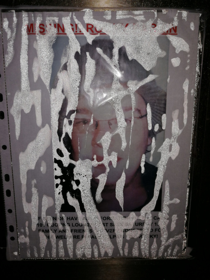 Partially obscured missing person poster. The poster features a photograph of a person with glasses, wearing a hat. The photo is heavily vandalised or defaced with white splotches and streaks, obscuring much of the person's face and the text below the photo. The visible text indicates the individual is missing and includes contact information, although much of it is illegible due to the damage. The overall effect is one of decay and despair, highlighting both the missing person's vulnerability and the indifference or hostility of the act of vandalism. The image’s story is one of loss, potentially compounded by the disrespect shown through the defacement of the poster, emphasizing the vulnerability of the missing person and the helplessness of those seeking them. The moral implication is a commentary on the fragility of life, the importance of community support in times of crisis, and also perhaps a reflection on urban decay and societal apathy.