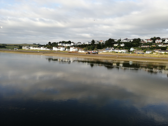 Tranquil waterfront scene. A calm body of water reflects a cloudy sky and the buildings of a small town or village situated on a hillside in the background. The foreground is a relatively flat shoreline with a sandy or muddy texture. A few boats or barges are visible near the shore.
