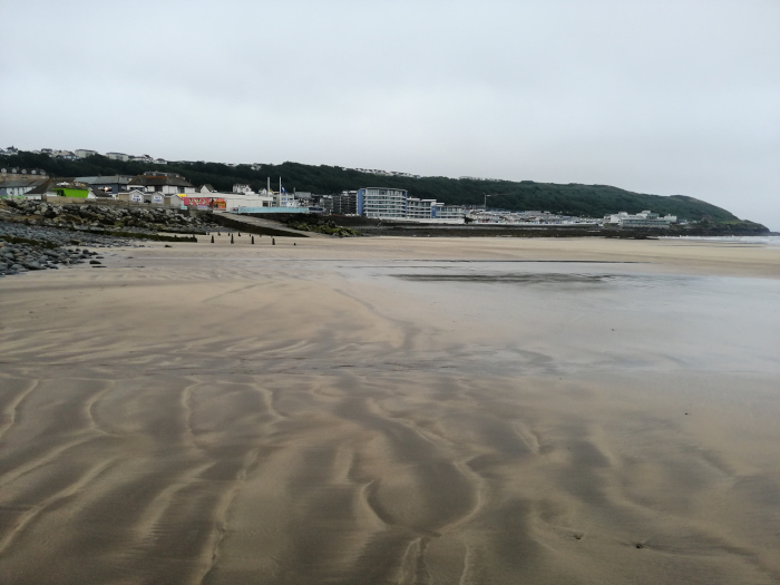 Wide sandy beach at low tide, with the receding water leaving behind intricate patterns in the sand. In the background, a line of buildings, including hotels and possibly shops, sits along the coast, nestled against a hill covered in green vegetation. 
