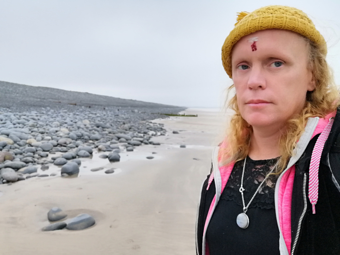 Leonie standing on a beach. She's wearing a yellow knit hat and a pink jacket. The beach is mostly sandy with a line of large, grey stones along the water's edge. The sky is overcast. There's a small, possibly symbolic, red mark on her forehead. She is directly facing the camera with a serious expression.