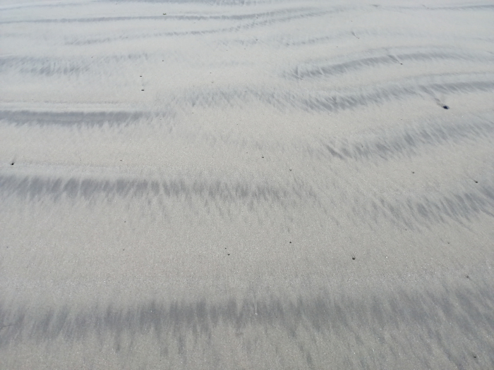 Close-up view of a sandy beach. The sand is light gray with darker gray streaks and patterns created by the wind or water. The texture is smooth but shows the ripples and undulations caused by the movement of the sand. There are a few small, dark specks scattered across the surface. The overall impression is one of simplicity and natural texture.