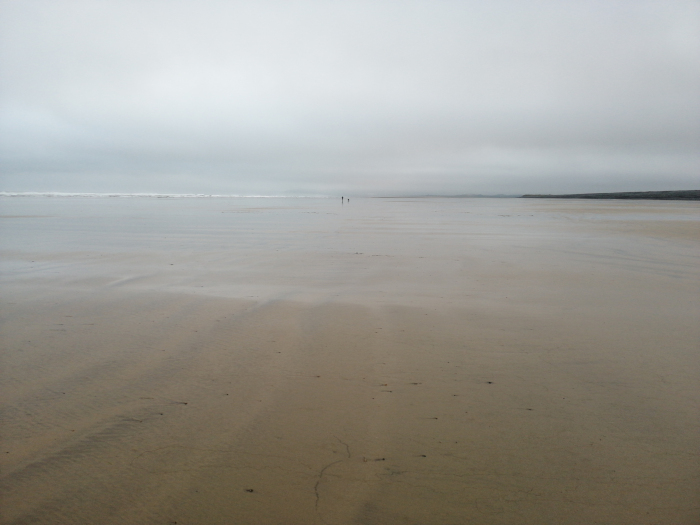 Wide, expansive beach scene under a muted, overcast sky. The sand is a light beige, wet in places, and stretches to the horizon where a faint line of ocean meets the sky. Two tiny, distant figures are barely visible near the waterline, suggesting vastness and solitude. The overall mood is calm, quiet, and somewhat melancholic due to the grey tones dominating the image.