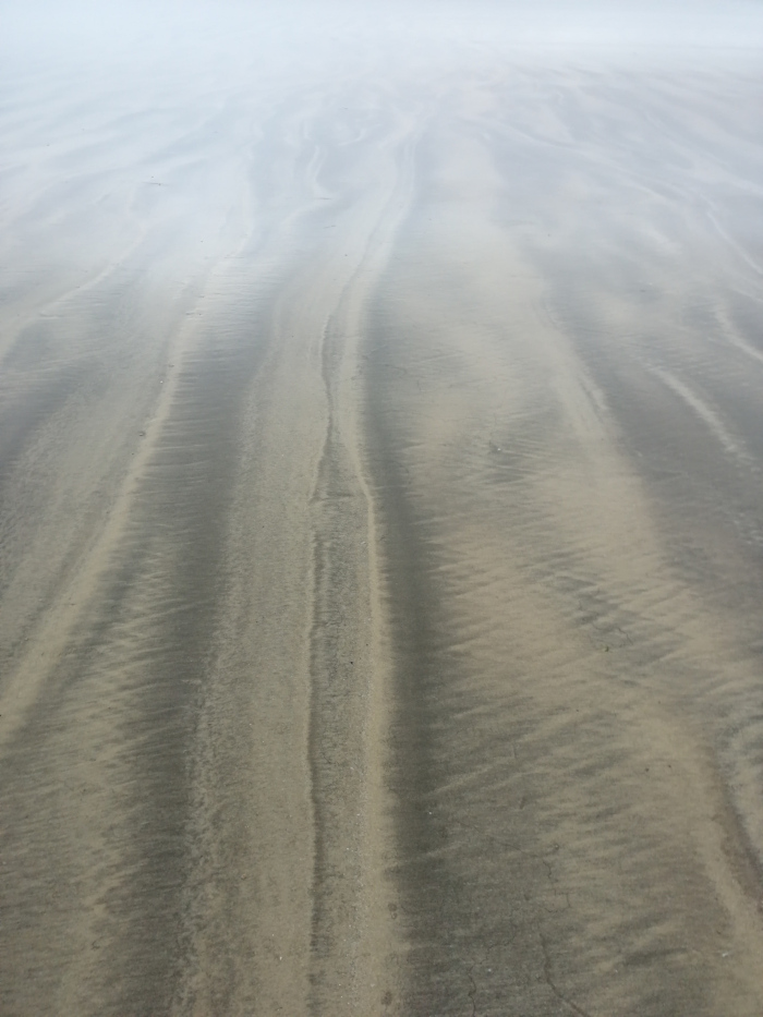 Beach scene with wet sand exhibiting patterns created by the receding tide. The lines in the sand are predominantly dark gray and lighter beige, running parallel to each other, creating a textured effect. A light fog or mist hangs in the air, softening the overall appearance. The perspective is from directly above, looking down at the sand.