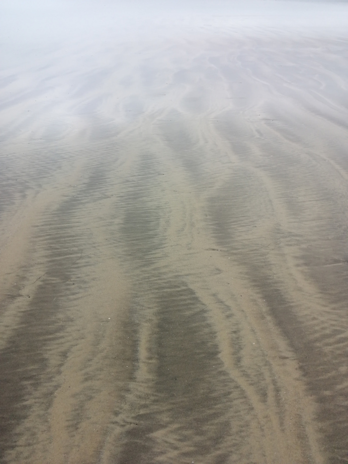 Beach scene, specifically the wet sand patterned by the receding tide. The patterns are a series of flowing, curving lines in light and dark shades of beige and grey. A light fog or mist hangs in the air, obscuring the background.