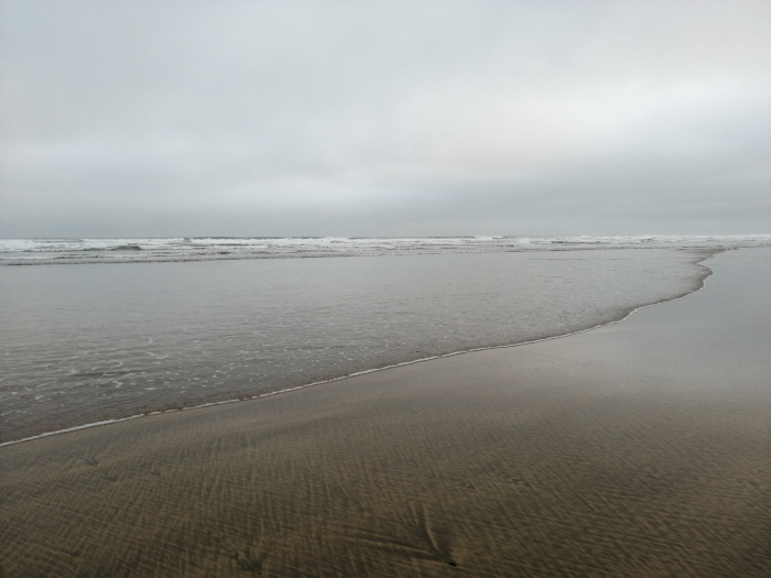 Tranquil beach scene on an overcast day. The water is calm, with gentle waves lapping the shore. The sand is wet and shows patterns from receding water. The overall mood is serene and muted due to the grey sky and subdued colours.