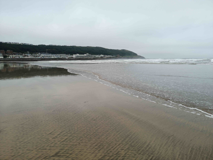 Wide, sandy beach with gentle waves lapping the shore. In the background, a line of buildings is visible against a low, green hill under a cloudy sky. The overall mood is serene and somewhat muted due to the overcast conditions. The sand shows patterns created by the receding tide.