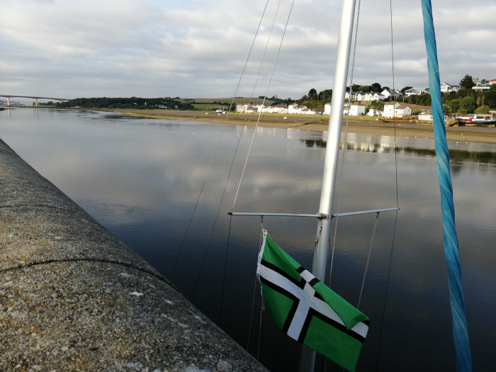 Calm river scene, viewed from a sailboat. A green and white flag, likely representing a region or county, is affixed to the sailboat's mast. In the background, a row of houses is visible on the riverbank, along with a bridge in the far distance. The overall mood is peaceful and serene. The focus is on the flag and the reflective water, creating a sense of place and possibly national or regional pride.