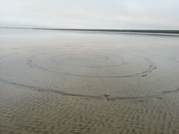 Large spiral pattern etched into the wet sand of a shallow body of water. The spiral is created by tire tracks from a vehicle that has repeatedly circled in concentric loops. The overall tone is muted and tranquil, with a flat, calm expanse of water extending to a distant shoreline under a cloudy sky.
