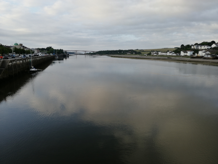 Calm river scene, likely at dawn or dusk, judging by the muted light. A bridge spans the river in the mid-ground, with a town on the left bank featuring buildings and parked cars. On the right bank, there's a line of houses. The water is relatively still, creating a mirror-like reflection of the cloudy sky. 
