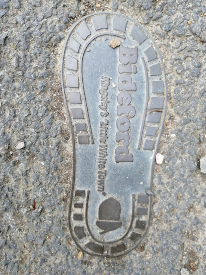 Metal plate embedded in the ground. The plate is shaped like a footprint and has the words Bideford Kingsley's Little White Town embossed on it. A small symbol, possibly a building or hat, is also visible at the bottom.
