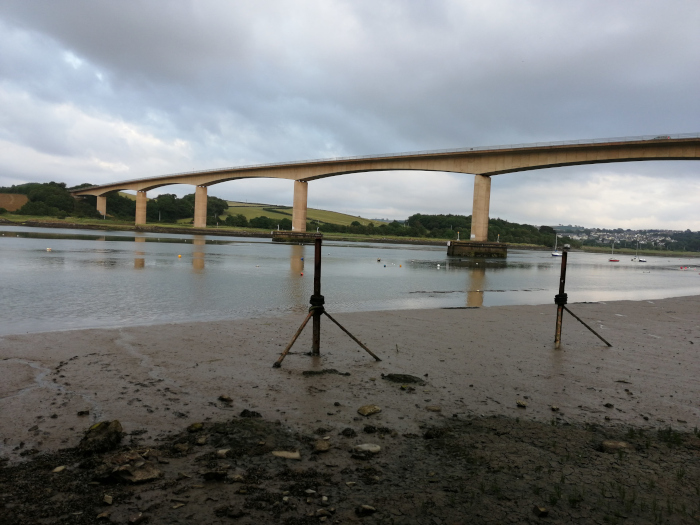 Long, high bridge extending across a calm body of water under a cloudy sky.  The foreground is a muddy, exposed shoreline with some rocks and two metal posts. A few small boats are visible in the distance on the water. The overall impression is one of a quiet, somewhat overcast day in a coastal or riverine setting.