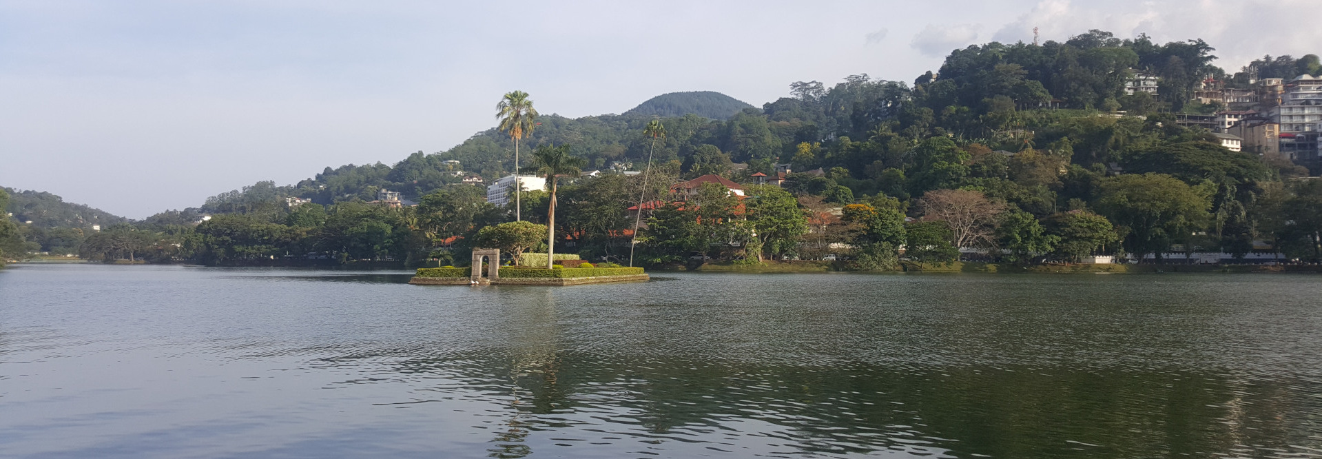 Lake's edge with a vibrant green hillside densely packed with lush tropical vegetation. Varied shades of green suggest different types of foliage; some trees appear fuller, darker, and others more sparse. The hillside slopes gently upwards into rolling hills forming a backdrop, culminating in a darker, more distant mountain under a pale sky. Buildings, appearing as light-coloured structures of various sizes nestled amongst the green, populate the upper portion of the hillside, hinting at a town or village. The lighting in the image is soft and diffused, suggesting either an overcast day or soft morning/evening light. The overall colour palette is muted, with greens, greys, and subtle browns dominating. The absence of strong shadows and the gentle light enhance the peaceful and tranquil atmosphere. The composition is panoramic, offering a wide view of the lake and its surroundings. The perspective is taken from across the lake, giving a sense of calm and distance. The horizontal lines of the lake and the hillside create a sense of depth and stability. The slightly off-centre placement of the island subtly breaks the symmetry, directing the eye's path across the water. The overall atmosphere is one of serene tranquillity and peaceful beauty. The calmness of the lake, the richness of the vegetation, and the muted light evoke a feeling of quiet contemplation and escape. There is a sense of harmony between the natural elements and the human presence suggested by the buildings in the distance. The scene projects a peaceful and contemplative mood, ideal for reflection.
