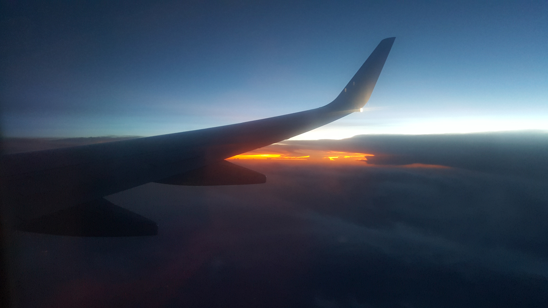 Portion of an air plane's wing, specifically the trailing edge and a section of the flap, sharply silhouetted against a vibrant sunset. The wing dominates the left side of the frame, angled slightly upward and away from the viewer. The wing is dark, almost black, with subtle variations in tone that hint at its three-dimensional form. There are no people visible. The wing appears to be in motion, carrying the plane through the air.