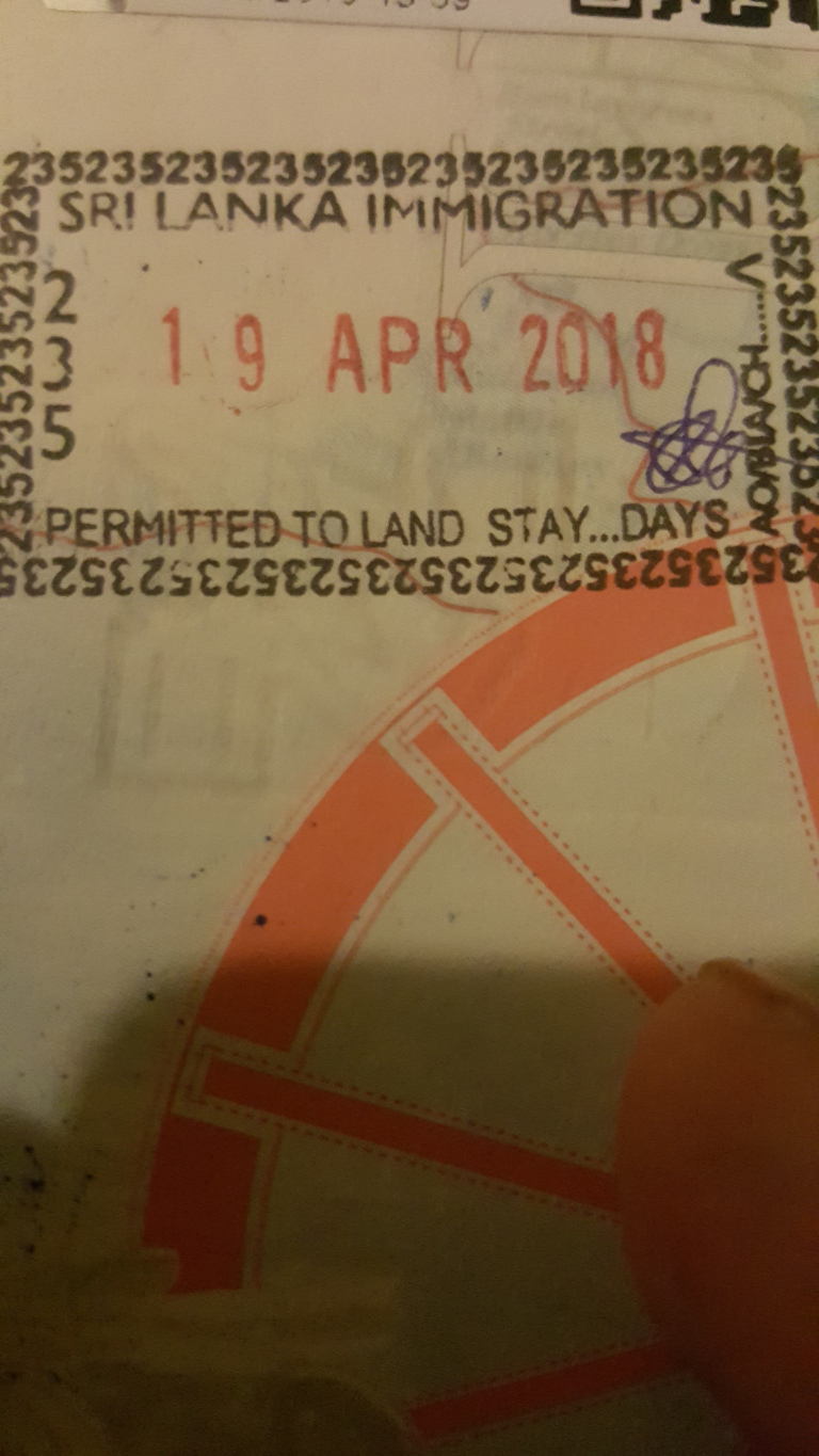 Close-up shot of a Sri Lankan immigration stamp affixed to what appears to be a passport or similar travel document. The stamp is the central focus, occupying a significant portion of the frame. The background is subtly visible, showing a continuation of the document's page, with faded markings and a faint, pale orange and white geometric pattern suggesting a design element of the document itself. This background fades into darkness towards the edges of the image, drawing attention to the central stamp.