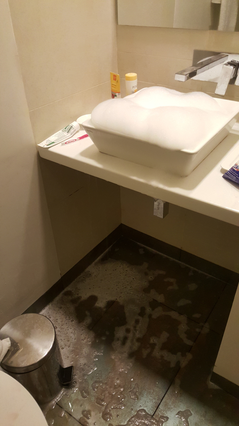 Bathroom scene dominated by a significant soap spillage. A square, off-white sink sits atop a matching counter top, overflowing with a generous mound of thick, white soap suds. These suds are billowing over the sides of the sink. On the counter top beside the sink are two small cosmetic containers — one yellow and one possibly pink or peach-coloured — suggesting toiletries. A partially visible toothpaste tube and what looks like a pink toothbrush lie to the left of the sink. The soap suds have clearly spilled onto the dark gray-brown floor, spreading across a considerable area. The suds are not uniformly distributed, creating a mottled pattern of wet, foamy patches. Under the sink is a small, stainless steel wastebasket. A portion of a toilet is visible in the bottom left corner.