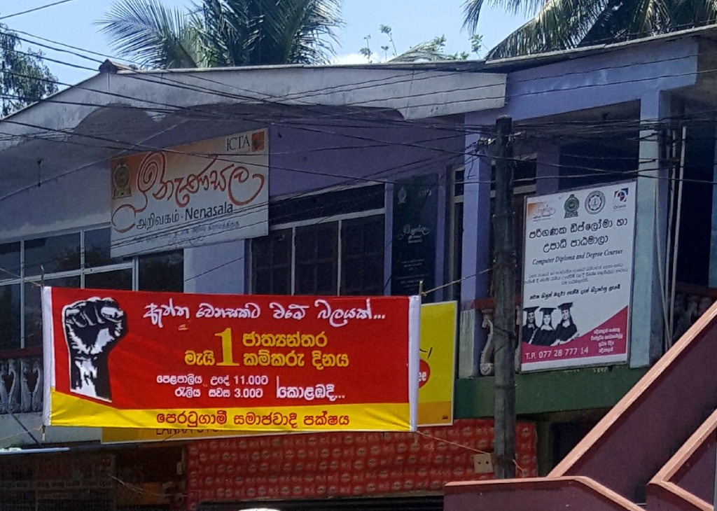 Two-story building, light purple in hue, with a slightly sloped roof. Several signs are prominently displayed. The most prominent is a large red banner hanging across the building's facade. It depicts a raised fist in a bold, graphic style, emblematic of protest or solidarity, against a predominantly red background with yellow accents. Text in Sinhala and Tamil is visible, advertising what seems to be a rally or demonstration, indicating dates and times. The numbers 1 and 11.000 stand out, possibly signifying a date and time, or a symbolic quantity. Beneath this banner are other smaller signs. One displays the name Nenasala in Tamil and Sinhala, suggesting a community centre or learning centre. Another sign advertises computer diploma and degree courses, with the contact details visible. Images of graduates are also present on this sign.