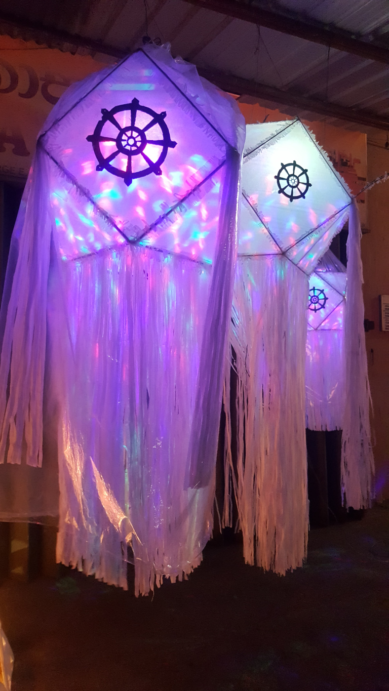 Two large, luminous lanterns hanging from the ceiling. They are diamond-shaped, constructed from a translucent white material, possibly plastic or a similar lightweight fabric. The material is slightly crumpled, giving a soft, uneven texture.  Each lantern features a dark silhouette of a Dharmachakra (the Buddhist Wheel of Law) prominently displayed in the centre. From the bottom edges of the diamond shapes, long, thin, white fringes hang down, creating a cascading, ethereal effect. The lanterns are illuminated from within with multicoloured LED lights, predominantly purples, pinks, and blues, which shine through the translucent material and are also reflected in the fringes, creating a shimmering, almost magical quality. The lanterns appear to be suspended from simple wire or string.