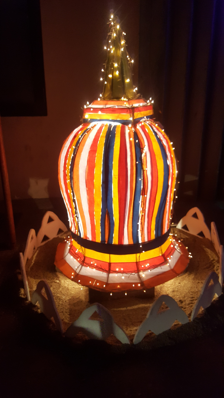Vertically oriented, multi-colored structure. The body is predominantly cylindrical, adorned with vertical stripes of orange, red, yellow, white, and dark blue, creating a bold, almost radiating pattern. The stripes appear to be made of fabric or paper, slightly translucent, allowing the internal lights to glow through. Atop the cylindrical body is a pointed, conical top, a darker yellowish-green, also wrapped in fairy lights. The base of the structure is wider than the body, giving it a slightly bulbous form. It rests on a sandy-coloured, circular platform, which is edged with stylised, low, white, mountain-like shapes arranged in a circle. There are small, warm-white LED lights woven throughout the structure, illuminating the colorful stripes and casting a soft glow around it.