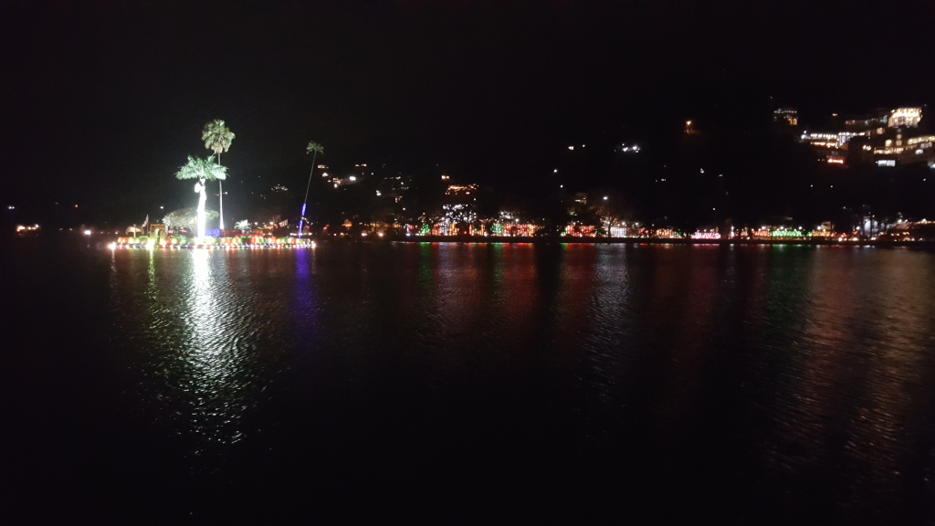 Nocturnal vista of a lakeside town, possibly in a tropical or subtropical climate, dramatically lit for a nighttime celebration or festival. The immediate foreground is dominated by the dark, still waters of a lake, reflecting the lights of the opposite shore like a shimmering, broken mirror. The reflections are fragmented, creating a mesmerising pattern of red, green, blue, and white light dancing on the water's surface. A small island or platform sits centrally in the middle ground, subtly illuminated. Two tall palm trees stand prominently on this platform, their fronds catching and diffusing the artificial light. The platform appears adorned with strings of lights, creating a festive aura around the palms. The background is filled with the silhouette of a hill or mountainside, densely populated with buildings. These buildings are indistinct in shape but are clearly lit from within, their windows glowing with various colours, creating a lively, continuous band of light along the horizon. The intensity of the lighting suggests the town is celebrating an event. No people are visible.