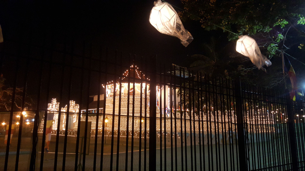 Large building, possibly a temple or palace, entirely encased in a warm, golden glow from countless small lights strung across its façade. The building's architecture is partially obscured by a tall, black metal fence running across the foreground. The fence's vertical bars create a rhythmic pattern that partially masks the building's details. Behind the fence, figures appear as indistinct silhouettes, seemingly moving, implying some kind of activity or procession in front of the illuminated structure. In the upper portion of the image, three translucent, lantern-like objects are suspended from wires strung between trees; they are illuminated from within, casting a soft, diffused glow. The scene is set at night, creating a strong contrast between the darkness and the brilliantly lit building. The sky is a deep, inky black, devoid of stars. Lush, dark foliage of trees is visible above and to the sides of the building, partially obscuring the background. The primary color is the warm, yellowish-white of the countless lights on the building, creating a festive ambiance. The lanterns hanging above add a soft, ethereal white light. The black metal fence offers a stark contrast against the warm lights.