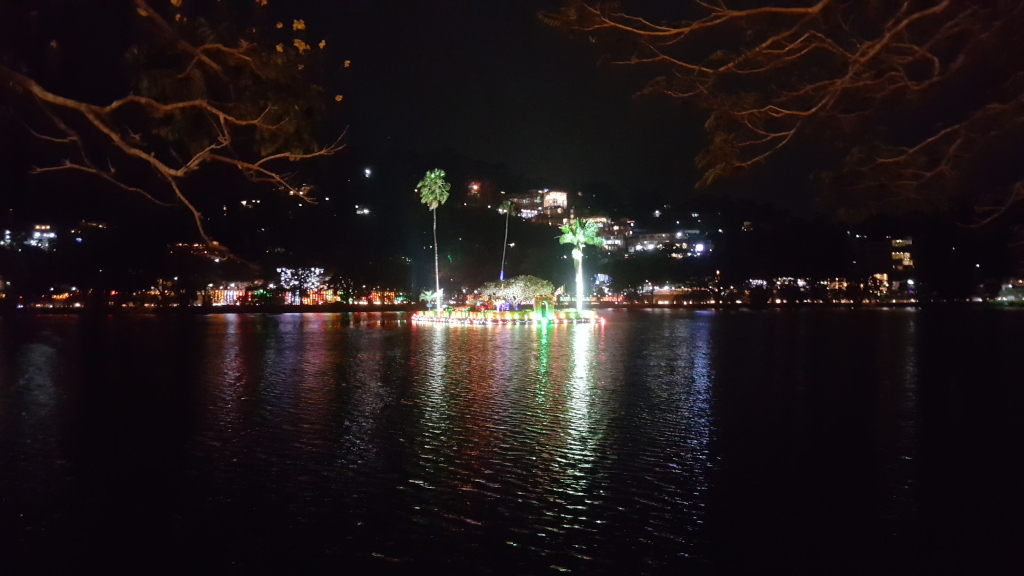 Nighttime scene of a lake, possibly Kandy Lake in Sri Lanka, with a city nestled on a hillside in the background. A dark, still body of water dominates the foreground, reflecting the lights from the city and a central illuminated island. The water's surface is gently rippled, indicating a calm atmosphere. In the middle of the lake is a small island, or possibly a floating structure, brilliantly lit with multicoloured lights – reds, greens, and whites are prominent. Several palm trees are visible on this island, adding a tropical touch.