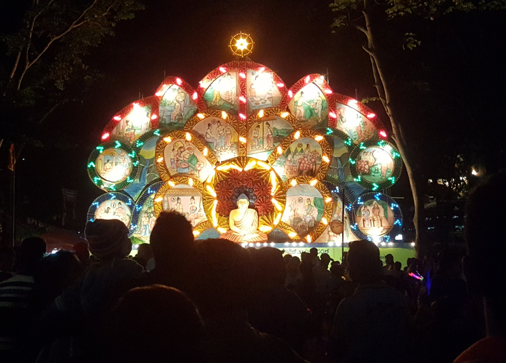 Large, illuminated display depicting scenes from the life of Buddha. The display is shaped like a stylised lotus flower with multiple circular panels, each showcasing a different event or scene. These panels are vibrant, with predominantly warm colours (reds, oranges, yellows) accented by cooler greens and blues. The panels are framed by intricate, glowing beading or ornamentation. In the centre of the lotus, a large, illuminated image of Buddha sits in meditation. The Buddha is rendered in calm, peaceful pose, in warmer tones that complement the surrounding scenes. The display is surrounded by a crowd of people; their faces are mostly obscured by shadows and low lighting, but they appear to be observing the illuminated display. Many are dressed casually; only a few discernible details of clothing are visible due to the low light. A few individuals wear hats or head coverings. The crowd is tightly packed in front of the display, their heads forming a dark, undulating mass that frames the bright artwork. They are passive observers, seemingly absorbed by the spectacle.