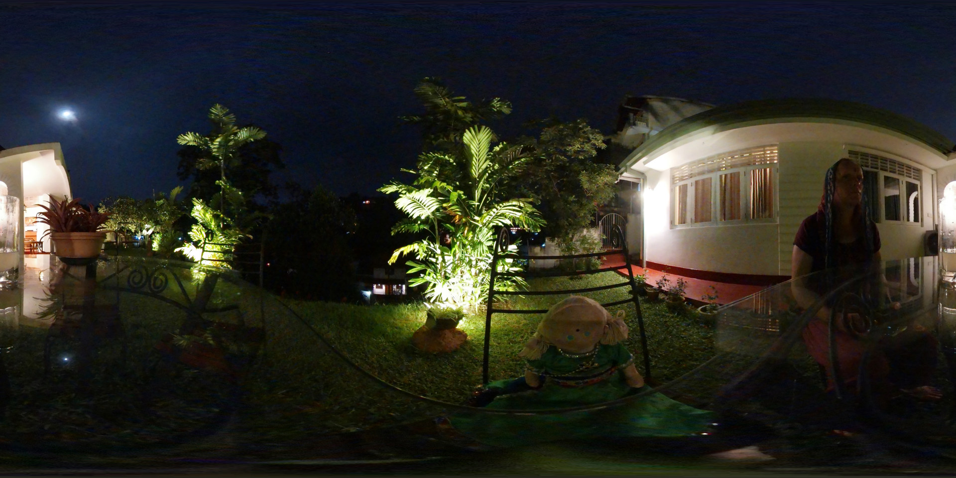 Nighttime scene in a garden. A person with fair skin, wearing a maroon top and a long, light-blue and white scarf draped over their head and shoulders, sits at a glass-topped table. They appear relaxed, almost contemplative, their hands resting on the table. Their posture suggests stillness and quiet observation. Near them, on the grass, sits a handcrafted doll with a light brown yarn head, painted features, a green dress with gold accents, and dark hair made of yarn. The doll is positioned slightly lower than the person, almost as if in a child-like pose. A potted plant is visible to the left, near a house. The setting is an outdoor garden area at night, illuminated primarily by artificial lights. Warm-toned lights highlight plants, particularly a palm-like tree, making its foliage glow. The lawn is a dark green, contrasting with the lighter-coloured house. The house is a colonial-style structure, white with a reddish-brown base, featuring large windows with visible curtains. The background reveals more vegetation, hinting at a hillside or sloped landscape. A faint moon is visible in the dark, slightly hazy night sky.