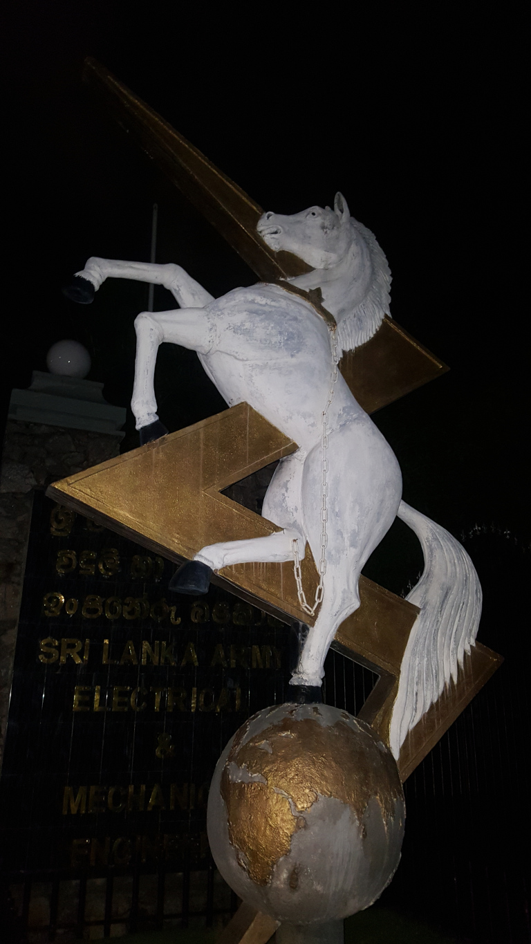Striking sculptural piece depicting a rearing white horse, seemingly powerful and dynamic. The horse is depicted mid-rear, its forelegs braced against a large, gold-coloured, lightning bolt-shaped structure. A chain runs from a collar around the horse's neck down to the base of the lightning bolt, implying restraint or perhaps harnessing of its energy. The horse is rendered with a slightly weathered or distressed paint job, the white showing some subtle textural variations. The lightning bolt itself has a slightly rough texture, the gold paint appearing possibly aged or patinated. Below the horse and lightning bolt, a sizeable, globe-like sphere rests, painted in muted gold and grey tones to suggest a terrestrial map. The sphere is somewhat rough in texture, showing variations in tone and apparent materially.