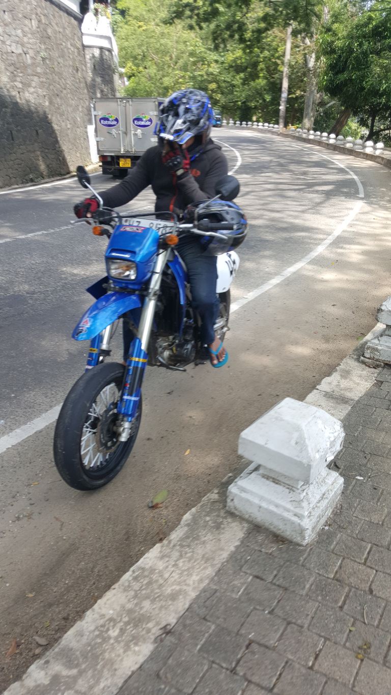 Person wearing a dark gray-black jacket, blue jeans, and bright teal flip-flops, riding a vibrant blue dual-sport motorcycle. The rider's head is mostly obscured by a dark-gray and white graphic helmet, partially covering the face. The rider's hands are visible gripping the handlebars, one hand appearing to hold a phone or device up to their face. They're positioned slightly off-centre, closer to the left side of the frame, moving along a winding road. The motorcycle is a Suzuki DR-Z400SM (based on the visible markings on the bike), with a blue body, white accents, and black wheels. The vehicle’s front wheel is distinctly in the foreground and the rear is slightly out of focus, suggesting a sense of movement. A smaller helmet is attached to the rear of the motorcycle. The setting is an outdoor scene on a curved road, flanked by a low stone wall with white concrete posts at regular intervals. Beyond the wall, lush green vegetation, including trees and bushes, is visible, suggesting a hilly or mountainous region. A white refrigerated truck with blue lettering (possibly a delivery vehicle, partially visible) is in the background, further along the road. The lighting suggests daytime, with natural sunlight casting shadows, indicating the scene likely occurred in a sunny climate.