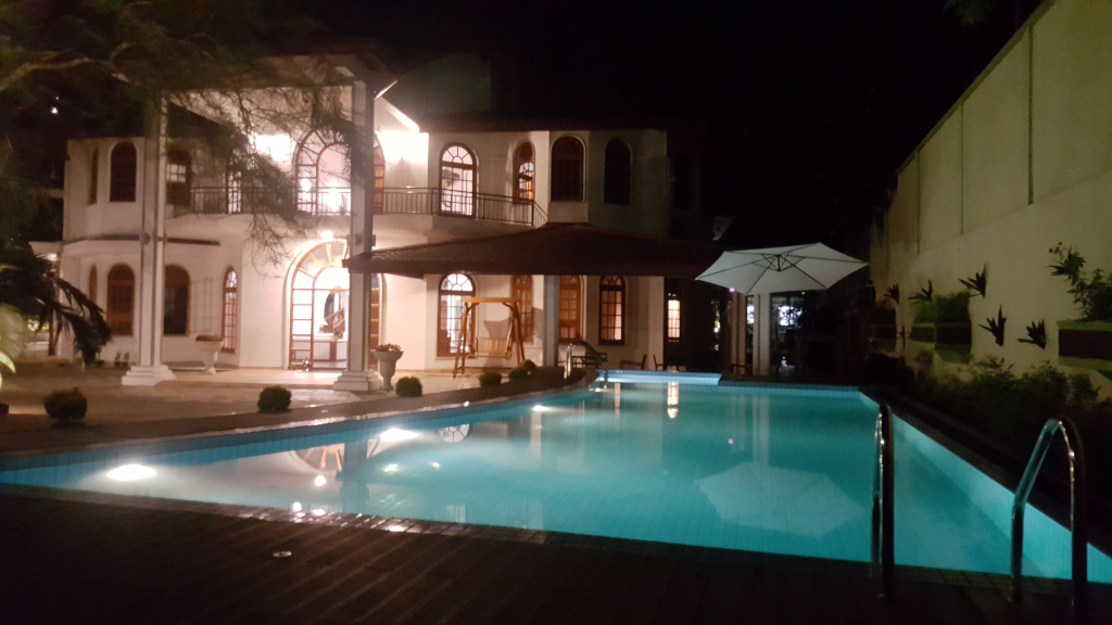Large, inviting swimming pool, its turquoise water reflecting the ambient lighting. The pool's shape is slightly kidney-shaped, curving gently away from the viewer. Stainless steel ladders are visible at the pool's edges. The pool is surrounded by a light-colored wooden deck. Behind the pool, a two-story white villa commands attention. The villa is brightly lit, showcasing its architectural details such as arched windows and a covered patio area with a wooden swing. Near the villa, under a beige-coloured patio cover, a wooden swing hangs gently. A white patio umbrella stands near the pool’s edge.  Lush greenery, including shrubs and trees, is sparsely visible around the perimeter of the pool and house. The scene is set at night. The predominant colours are the deep blue-green of the pool, contrasted by the bright white of the villa and the darker greens of the surrounding vegetation. The lighting is artificial, emanating mainly from the villa’s interior and exterior lighting fixtures, creating a warm, inviting glow. The background is mostly dark, with a suggestion of a tall wall to the right of the image. The overall lighting contributes to a sense of peaceful serenity. The colour palette is muted and calming, with the cool tones of the night contrasting with the warmer tones of the house's lighting.