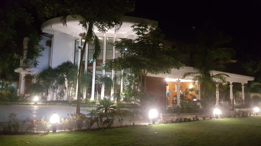 Large, modern building illuminated at night. The building features a striking white, curved façade with prominent glass windows spanning a significant portion of the structure's front. A lighter-colored, possibly brick or stucco, section extends from this main area. The building has a portico supported by white pillars. Inside, a dimly lit interior is visible through the glass, hinting at furniture and possibly a reception area. A person, seemingly a staff member in a light-colored shirt, is faintly visible inside the portico area. Landscaping surrounds the building, including lush tropical plants, palm trees, and neatly trimmed shrubs. Several low-level outdoor lights are strategically placed, casting a warm, inviting glow on the pathways and vegetation.