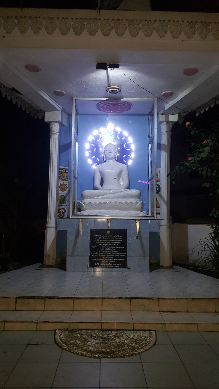 Serene, nighttime view of a small, white Buddha statue enshrined within a glass enclosure.A pristine white, seated Buddha statue dominates the scene. It's positioned in the middle of the image, within a glass case, with a lotus-like base. The statue is illuminated from behind by a circular halo of bright, cool-toned LED lights.