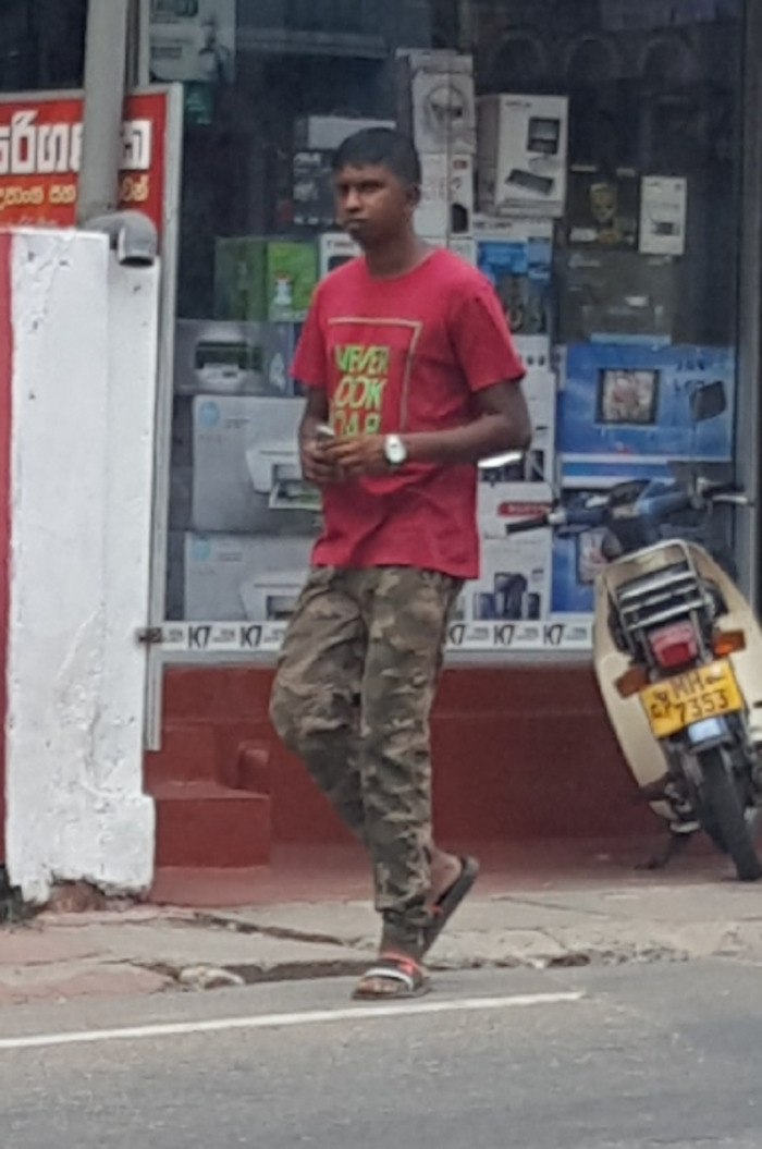 Young, slender man of South Asian descent, appearing to be in his late teens or early twenties. He's walking on a street, slightly angled towards the viewer's left. He wears a vibrant red t-shirt with a partially visible lime green and white graphic design that seems to contain words. His pants are camouflage-patterned in muted greens and browns. He's wearing simple, dark-coloured sandals, one of which is clearly visible while the other is partially obscured. He carries something small in his left hand, possibly a phone, and a watch is visible on his left wrist. His expression is neutral, almost pensive. To the right of the man is a pale beige-colored motor scooter, parked, facing towards the viewer's left. Its license plate is clearly visible.