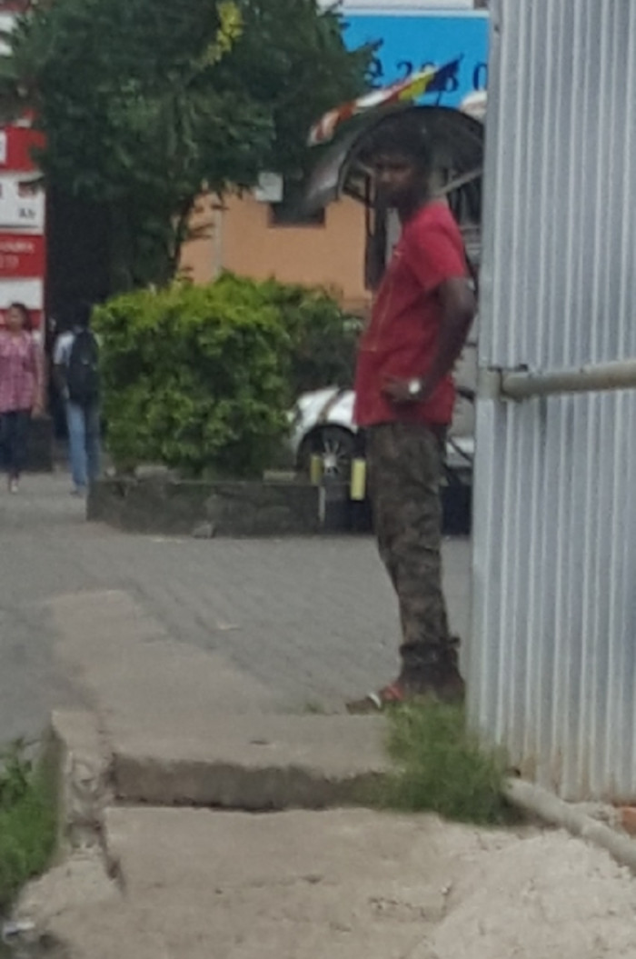 Man, appearing to be of South Asian descent, standing slightly angled to the right, partially obscured by a corrugated metal fence. He's wearing a vibrant crimson short-sleeved t-shirt and camouflage-patterned trousers, tucked into sturdy, dark brown boots. His posture is relaxed but attentive, his hands resting casually – one at his waist, the other near his side. A colourful umbrella, possibly a traditional design, rests on his head, partially visible behind him. In the background, other individuals are visible at varying distances. A woman in a pink and white patterned top and dark trousers walks away from the camera, appearing near the edge of a building. There is a man in a light-colored shirt and dark trousers walking behind the main subject, also distant and out of focus.