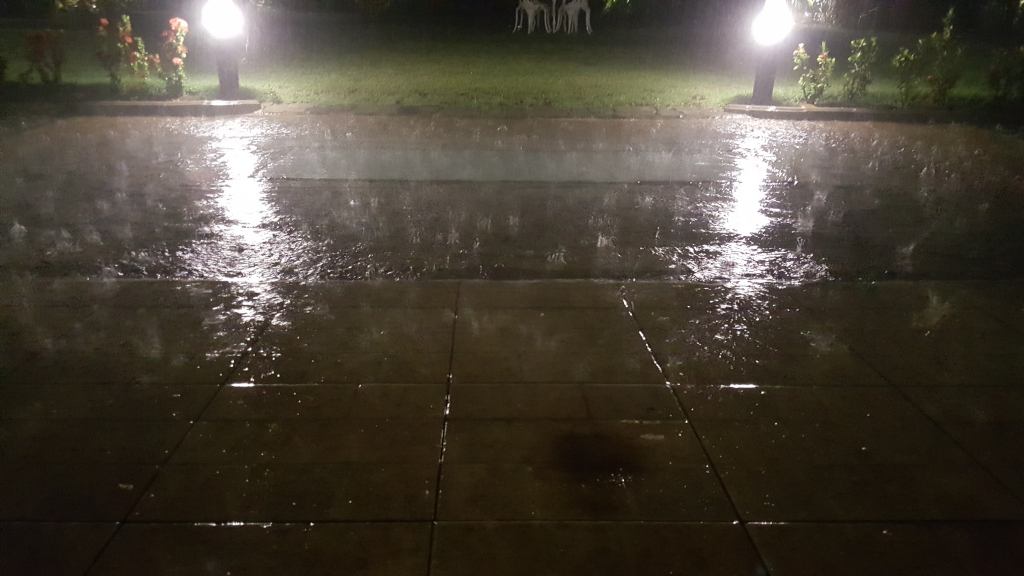 Paved surface, appearing to be a patio or walkway, glistening under a heavy nighttime downpour. Rain streaks across the tiles, creating small ripples and reflections of light. Two illuminated outdoor lamps stand at either end of the walkway, casting pools of bright light on the wet pavement. These pools of light are intensely bright and somewhat overexposed, creating a stark contrast with the surrounding darkness. The lamps are simple, upright cylindrical shapes with their light radiating downwards. In the background, just beyond the walkway, faintly illuminated, are indistinct shapes that appear to be small garden ornaments or statues.