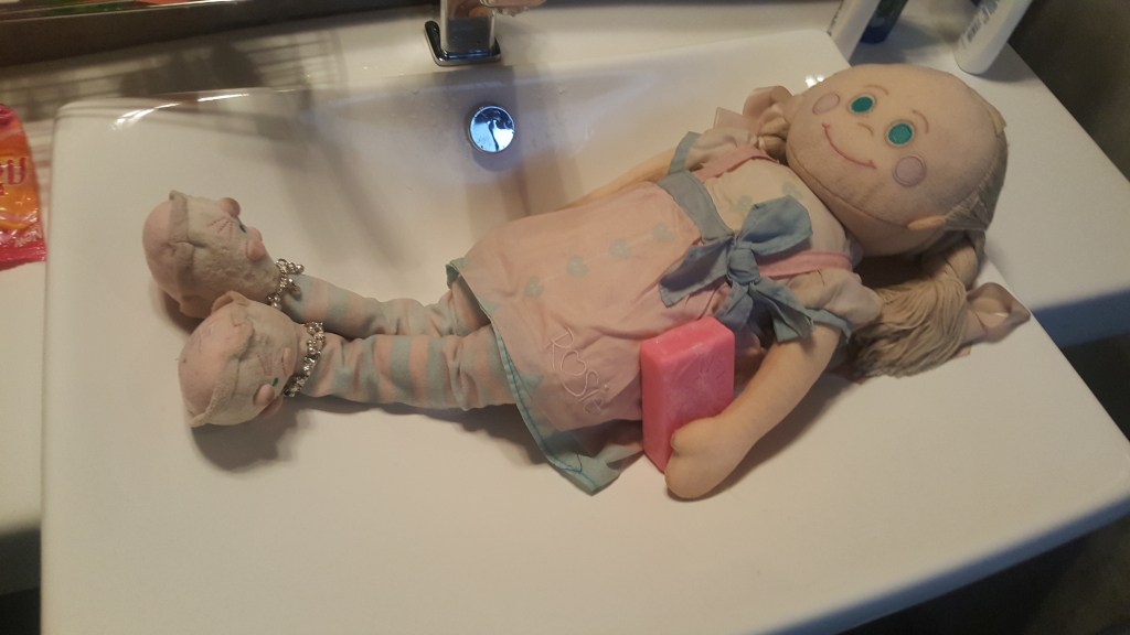 Rag doll, seemingly vintage or handmade, lying supine in a white bathroom sink. The doll is fair-skinned, with painted-on teal eyes and rosy cheeks. Its blonde hair is long, slightly unkempt and appears to be made of yarn or a similar material. It's wearing a pale pink and blue dress with a delicate light blue bow, and striped pastel blue and white leggings or stockings. The doll's feet are clad in miniature cloth slippers. The doll is positioned diagonally across the sink, arms relaxed at its sides, slightly curved, holding a small, rectangular, pale pink bar of soap against its midsection. Two smaller rag doll-like objects, possibly slippers or small dolls, are tucked beneath its legs.
