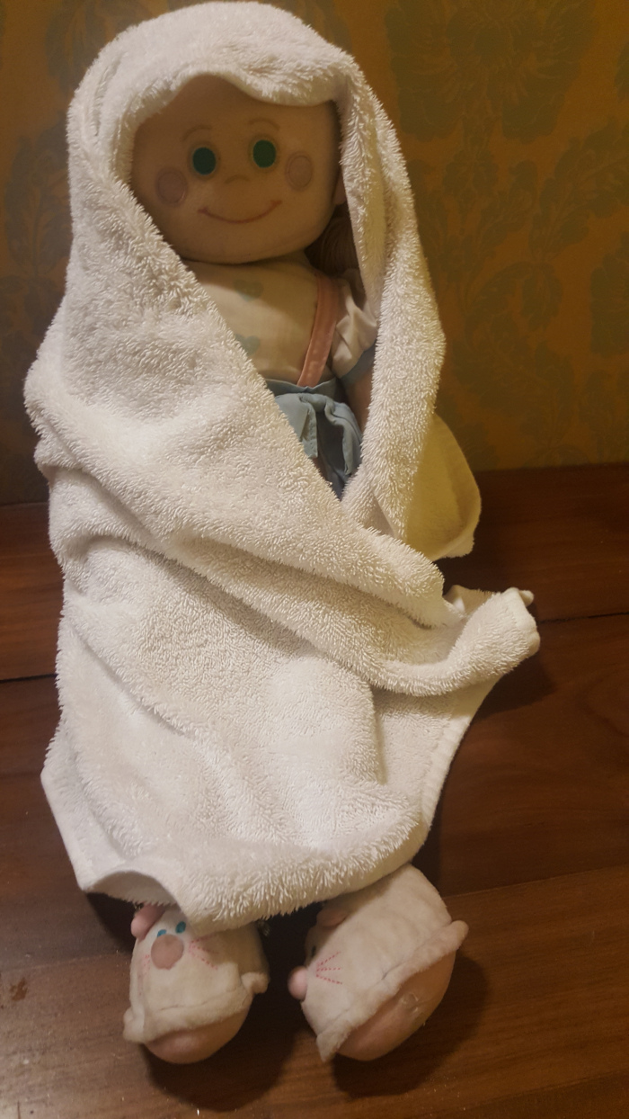 Rag doll, seemingly female, wrapped snugly in a large, fluffy, off-white bath towel. The doll's face is simple, with large, light green eyes, rosy cheeks, and a stitched-on smile. It wears a light-coloured, possibly pastel blue and white, outfit partially visible under the towel. The doll is seated, its legs tucked slightly under the towel, and it wears light beige or cream-colored slipper-like footwear with subtle animal-face designs (possibly mice or rabbits) embroidered on the toes. The doll appears peaceful and relaxed. The towel is generously sized, draped loosely yet securely around the doll, creating soft folds and textures. The scene is set against a warm, golden-brown wood surface which seems to be a table or floor. Behind the doll, the backdrop is a patterned wallpaper in muted yellowish-gold tones with a stylised floral or leaf design. The wallpaper's pattern is somewhat indistinct but contributes to a calm, domestic setting. The lighting is soft and diffused, preventing harsh shadows and casting a gentle glow over the doll.