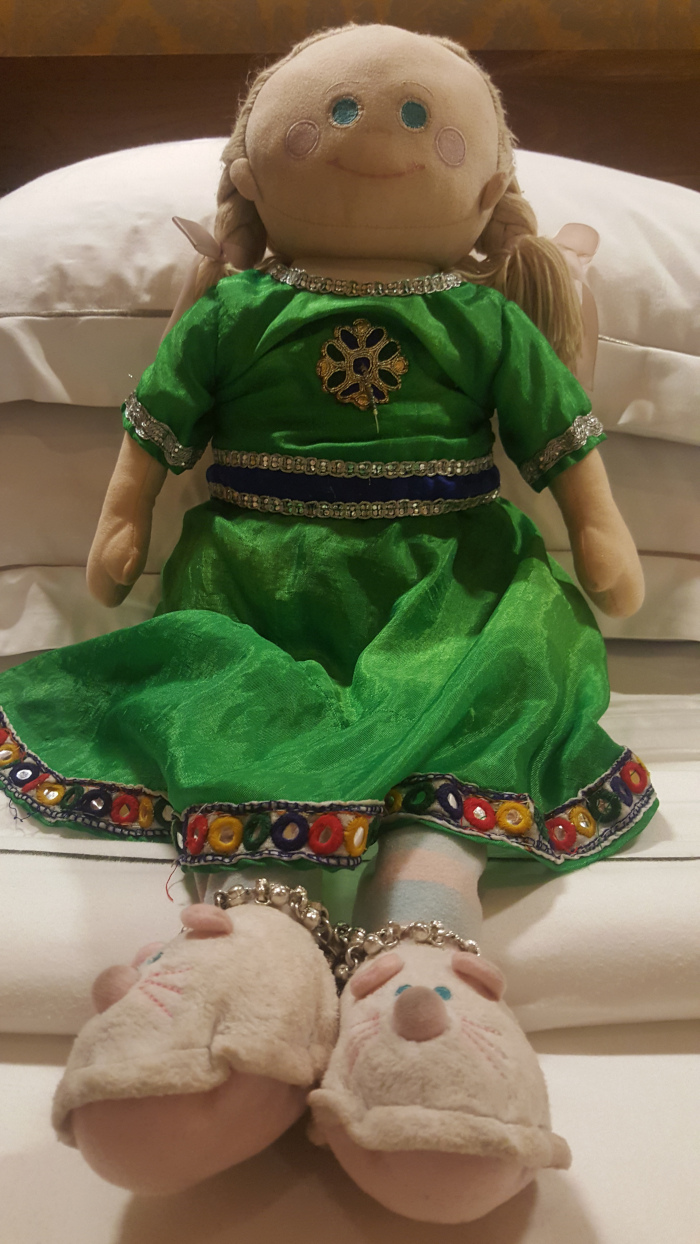 Rag doll, seated on a pristine white pillow. The doll is characterised by a light beige, almost creamy, face with simple, hand-stitched features: two slightly mismatched light blue eyes, rosy pink cheeks, and a subtle, serene smile. Its head is bald except for two pigtails of light blond yarn. The doll wears a vibrant green, flowing dress adorned with intricate details: a gold and blue floral emblem at the bodice, a navy blue and silver beaded belt, and a colourful, embroidered hemline showcasing red, yellow, green, and blue circles and mirrored embellishments. The doll's lower limbs are clad in pastel-striped socks and fluffy, off-white mouse-shaped slippers, each with tiny pink ears and blue eyes. A silver beaded anklet adorns each leg. The doll appears to be passively sitting, its expression placid and peaceful.
