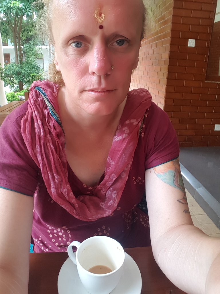 Leonie with shoulder-length, light blonde hair. Her expression is serious, almost melancholic; her gaze is directed slightly downward. She's seated at a dark brown wooden table, her body angled slightly towards the viewer. Her arms are visible, revealing a colorful bird tattoo on her right upper arm and several noticeable, pale scars along her left forearm. She wears a deep maroon, patterned dress with a sheer, reddish-pink scarf draped loosely around her neck. A small, gold-coloured bindi adorns her forehead, and a small nose stud is visible. She holds a small, white teacup with a light brown liquid (presumably tea) inside. The teacup sits on a white saucer.