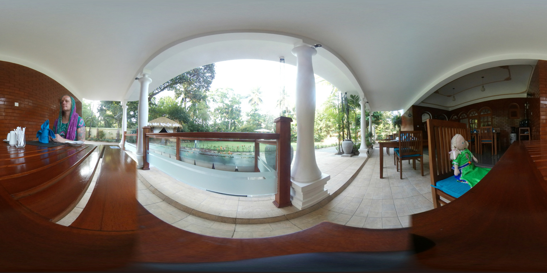 360-degree view of an outdoor eating area. Leonie with short hair, a tattoo visible on her left arm, and a patterned purple and teal headscarf is seated at a dark, polished wooden table. She appears calm and is sitting upright, seemingly waiting or observing something. She has a serious expression. A handmade-looking doll, dressed in a bright green and teal outfit, sits alone on a teal-cushioned chair at another table, further back in the scene. Several dark brown wooden tables and matching chairs with teal cushions are arranged in the dining area, both under cover and slightly visible beyond the partially visible glass enclosure.