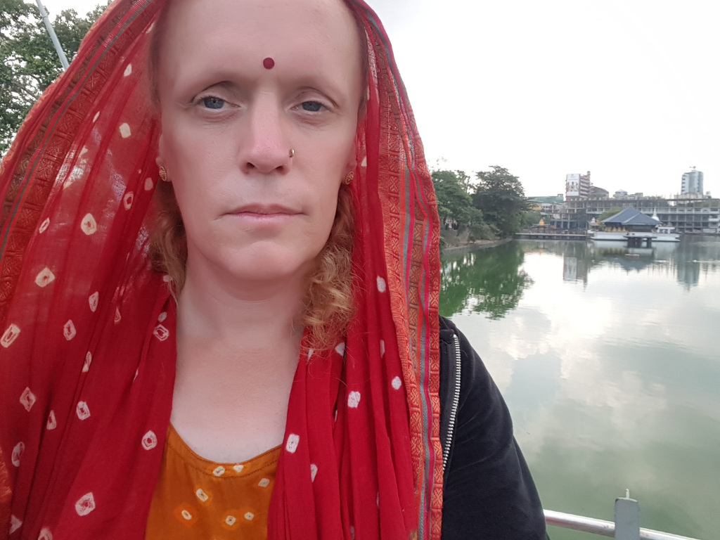 Person, seemingly androgynous, with fair skin, and a calm, somewhat serious expression. Their eyebrows are subtly arched, and they have a small, dark bindi (a red dot) centred on their forehead. They possess a nose piercing and small, gold-toned earrings are visible. Their hair is a light reddish-blonde, partially visible beneath a vibrant red and white patterned scarf, which is draped over their head and shoulders, The scarf is a lightweight fabric, appearing semi-transparent in places, revealing the person's clothing underneath – an orange-gold top or dress adorned with similar but smaller white patterns as the scarf. A black zip-up jacket is partially visible on their shoulders. The person appears to be standing, slightly angled, looking directly at the camera. There are no discernible actions beyond their pose.