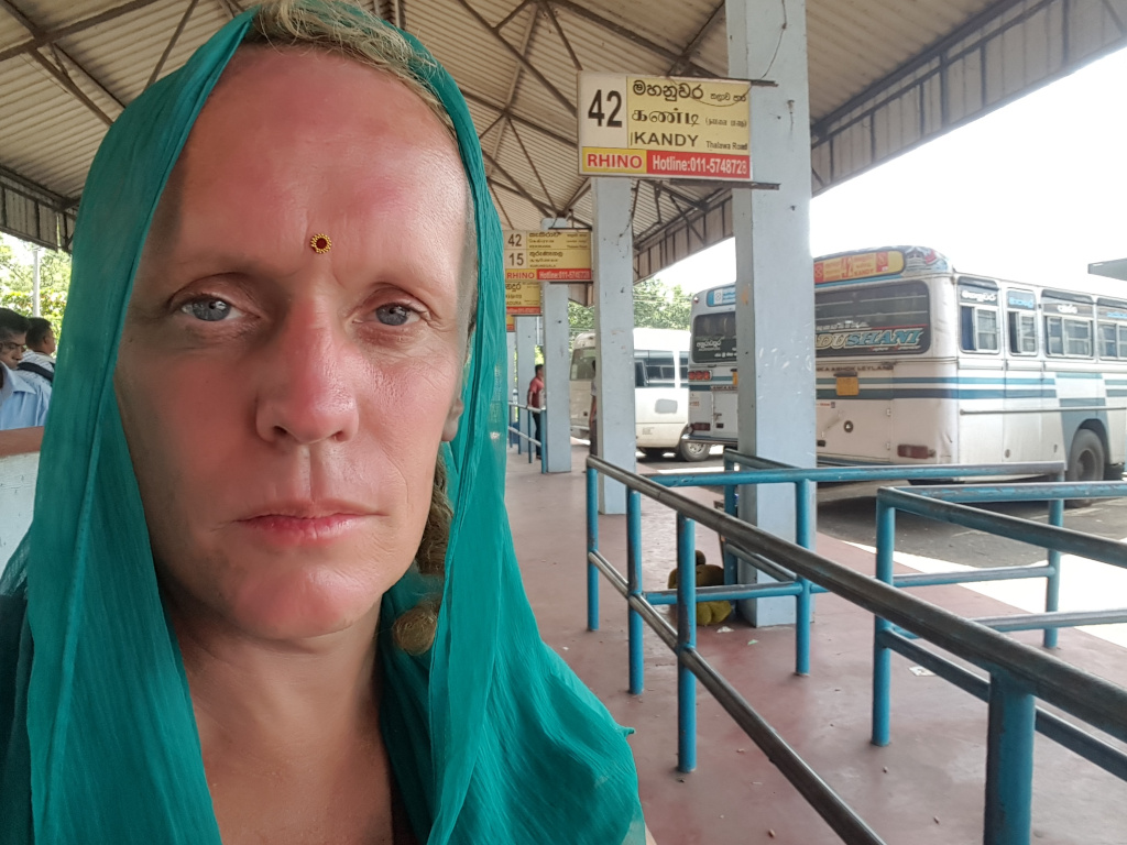 Fair-skinned woman, centrally positioned, who appears to be of Caucasian descent. She is wearing a vibrant teal-coloured headscarf, draped elegantly around her head and shoulders, concealing her hair and falling down to her chest. A small, round, reddish-orange bindi adorns her forehead between her eyebrows. Her expression is serious, perhaps pensive or weary, with her eyes looking directly at the camera. Her skin shows some signs of sun exposure or natural tan. In the background, several Sri Lankan buses are visible, parked at a bus stop. One bus is prominently featured, in focus behind the woman, displaying Sinhala and English script on its exterior. A child is partially visible, crouching behind a blue railing in the mid-ground. In the far left background, other individuals are partially visible, appearing to wait at the bus stop.