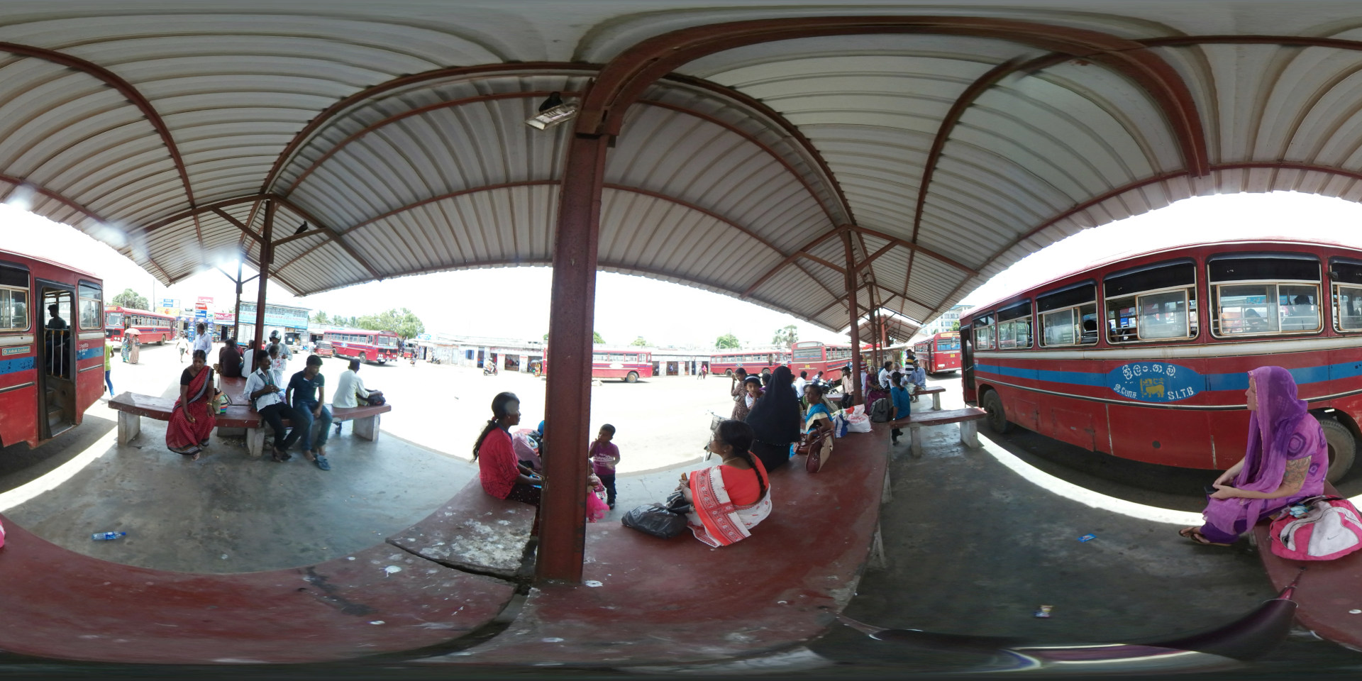 Bus station shelter, under which numerous people are seated or standing. The shelter itself is a low, wide structure with a corrugated metal roof curving dramatically downward to form a large, shaded area.  Several groups of people are visible, predominantly women and a few men. One woman in a vibrant crimson sari sits on a long bench, her posture relaxed. Near her, another woman in a maroon and white striped sari sits with her legs crossed, a serene expression on her face. Further along the bench, a group of three women sit, one wearing a deep purple sari, her arms folded, looking slightly pensive. Other individuals are scattered, some in small groups, engaging in casual conversation or simply waiting. A young child in a blue and white striped shirt is sitting near a group of women. The clothing is colourful, with many saris exhibiting a mixture of striking and earthy tones. A few men are seen in shirts and pants. One woman stands out, sitting near a red bus on the right, wearing a bright purple sari and what looks like a temporary tattoo on her arm. She is facing away from the other seated passengers, seemingly engrossed in thought.