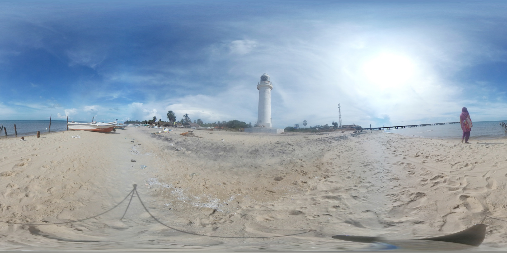 Slender, cylindrical white lighthouse stands prominently in the mid-ground, its top slightly off-centre towards the right. It's a stark white against the sandy beach and blue sky, appearing tall and somewhat solitary.  woman, dressed in a long, purplish-pink robe and headscarf, walks away from the viewer towards the right, along the beach line. She carries a small bag and appears to be walking alone, her figure small against the vastness of the landscape. Two weathered, white and orange fishing boats rest on the sand near the left edge of the frame, close to the waterline. They appear used, slightly tilting, indicating they haven’t been recently launched. The foreground and mid-ground are dominated by a wide expanse of pale, sandy beach. Footprints and scattered debris—including small pieces of plastic and other litter—are visible on the sand, suggesting human activity and a natural, slightly unkempt shoreline.
