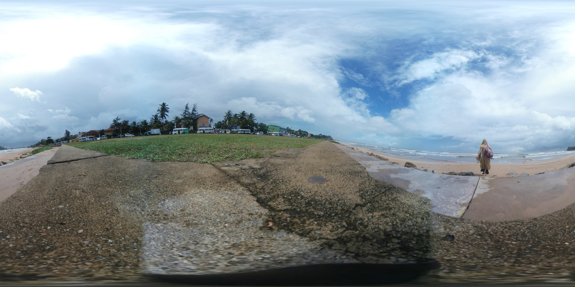 Leonie walking away from the camera towards the ocean. She's wearing a long, flowing, light-brown or beige dress and carries a maroon backpack. Her head is covered, possibly with a scarf or headscarf. The figure is positioned in the middle ground, providing a clear focal point within the panoramic view. The scene is a panoramic coastal view, captured at what appears to be a beachside promenade or seawall. The foreground is dominated by a rough, textured concrete seawall, showing signs of wear and tear with cracks and variations in color – primarily muted browns and grays, speckled with small stones and pebbles. Behind the seawall is a strip of short, green grass. Beyond the grass lies a line of buildings, including what looks like small hotels or guesthouses, interspersed with palm trees. The buildings are painted in light pastel colors. The background is the ocean, with light-colored sand at the shore and waves gently breaking. The sky is a vast expanse of a light blue, dotted with fluffy white cumulus clouds, which appear slightly hazy, suggesting either a morning or afternoon scene. The lighting seems natural and soft, with no harsh shadows.