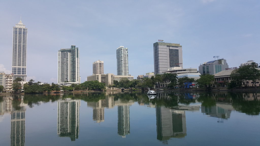 Picturesque cityscape reflected in a calm body of water, likely a lake or canal. The focal point is a row of modern high-rise buildings of varying heights and architectural styles, positioned along the far shoreline. The buildings are primarily light-coloured, a mix of off-white, beige, and light gray, with varying shades of glass windows. Some buildings have distinct architectural features, such as balconies or setbacks. Most prominent is a slender, light-colored skyscraper to the far left, standing taller than the others. To its right are several buildings of similar height, with one showing a darker glass façade. Further to the right is a slightly shorter, boxier building with a noticeable red sign near the top. The water is exceptionally still, creating a near-perfect mirror reflection of the cityscape and surrounding greenery. A small white boat is visible near the centre of the lake.
